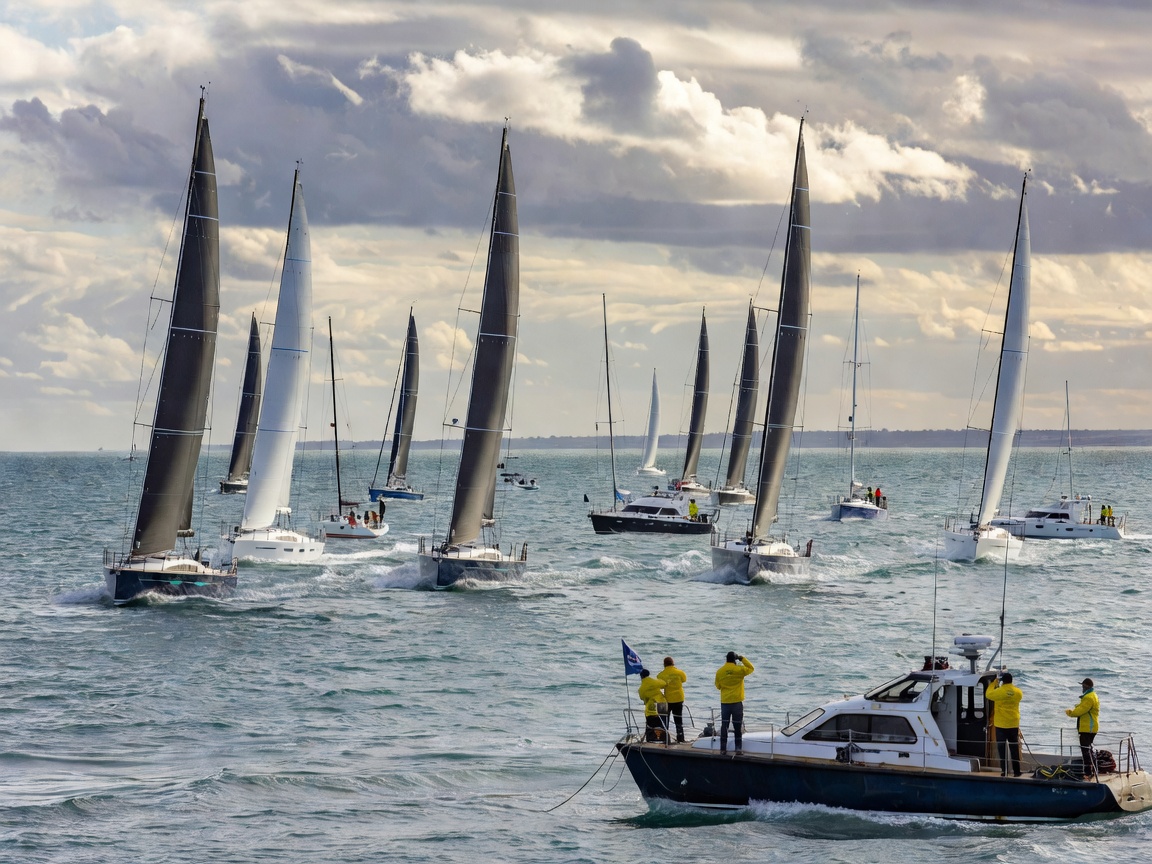 Fleet of racing yachts crossing a start line under full sail with a committee boat in the foreground